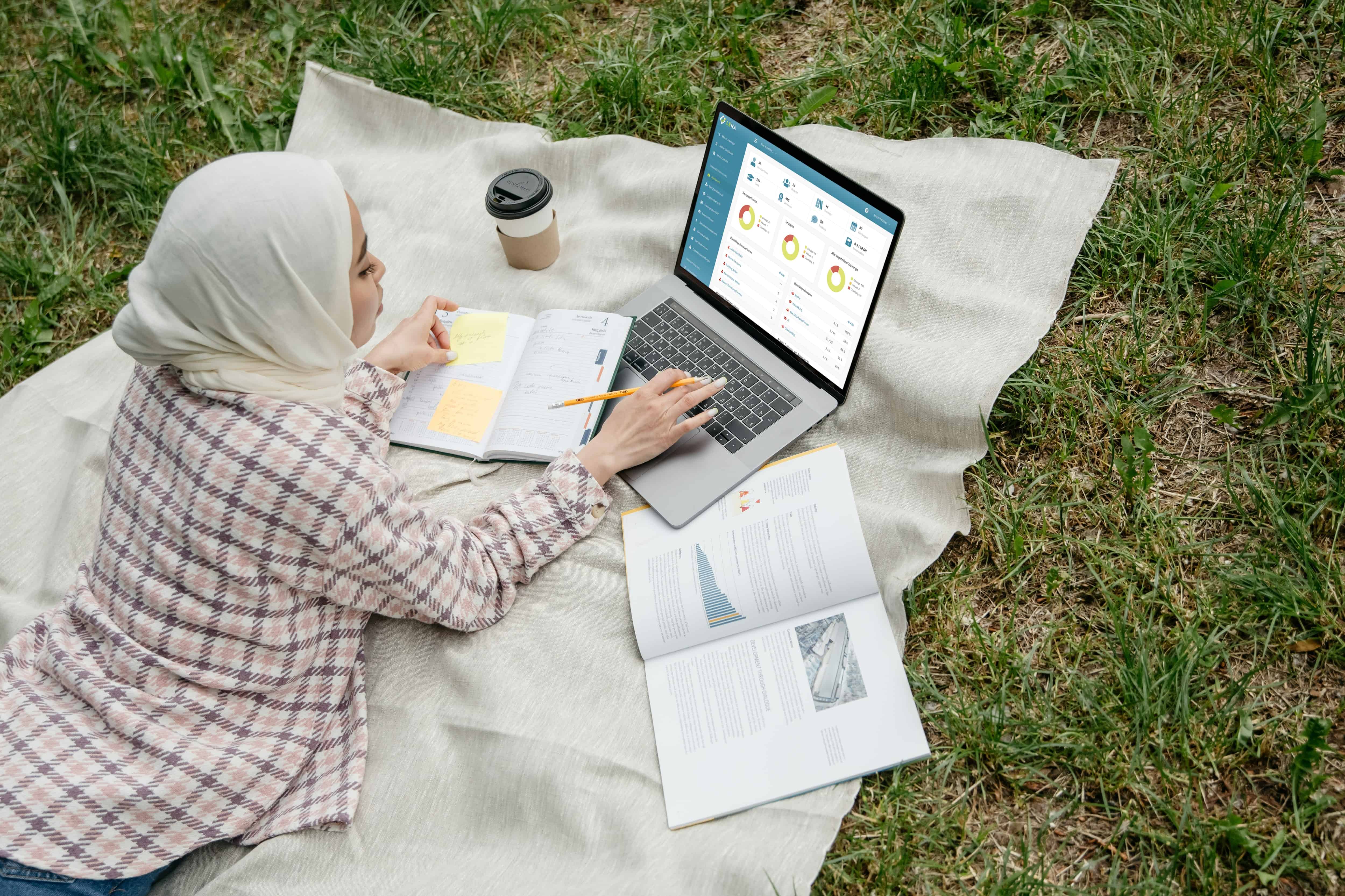 Frau liegt mit ihrem Laptop und Schulungsunterlagen auf einer Wiese und macht eine Online Schulung